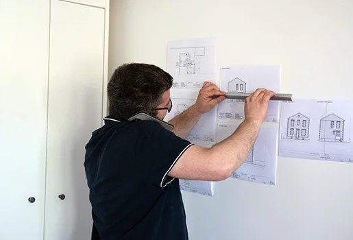 Architecte au téléphone mesurant des plans de maison affichés sur un mur à Cormeilles-en-Parisis dans le Val-d’Oise 95