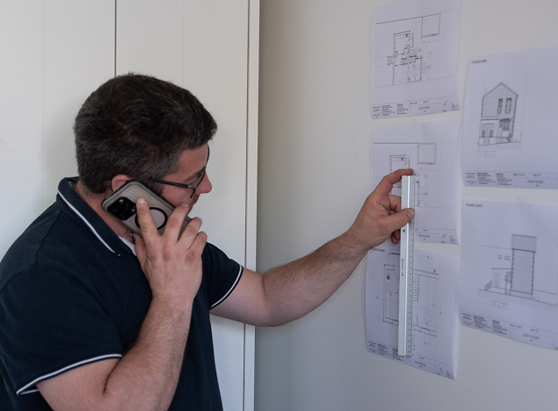 Homme au téléphone examinant des plans de maison sur un mur avec une règle à Cormeilles-en-Parisis dans le Val-d’Oise 95